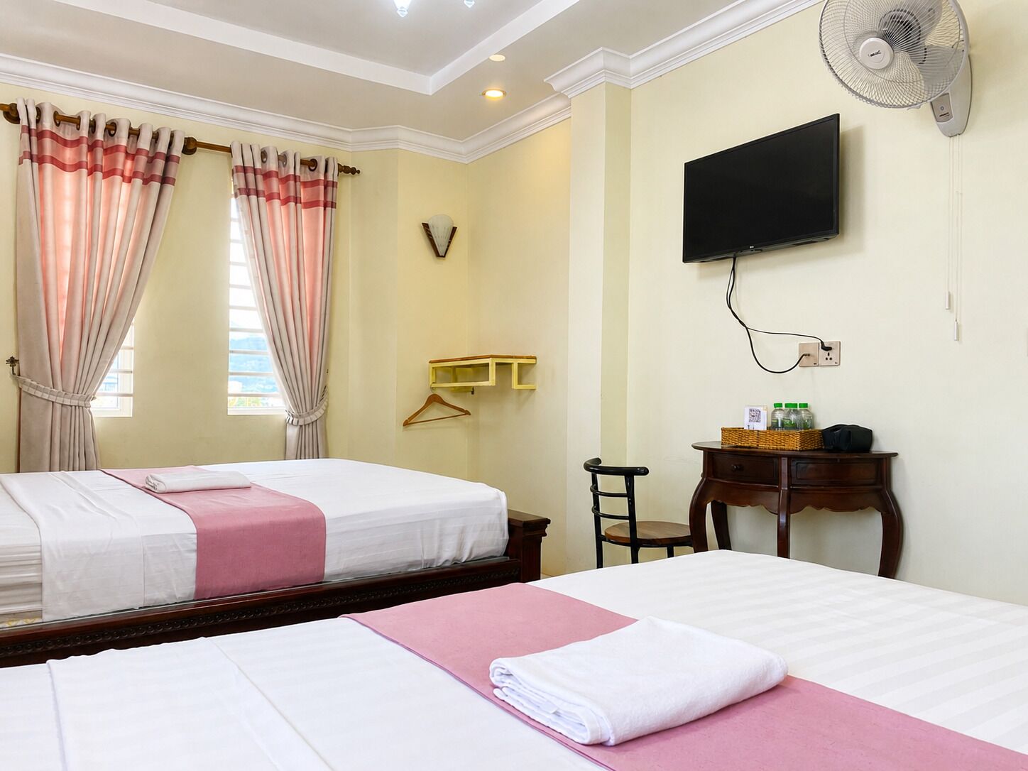 A hotel room with two beds, a television, a fan, and a small table with a basket and bottles.