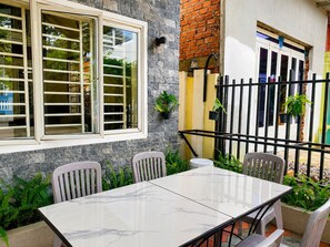 Un patio con una mesa y sillas de mármol, una ventana con persianas, un muro de ladrillo y plantas en macetas.