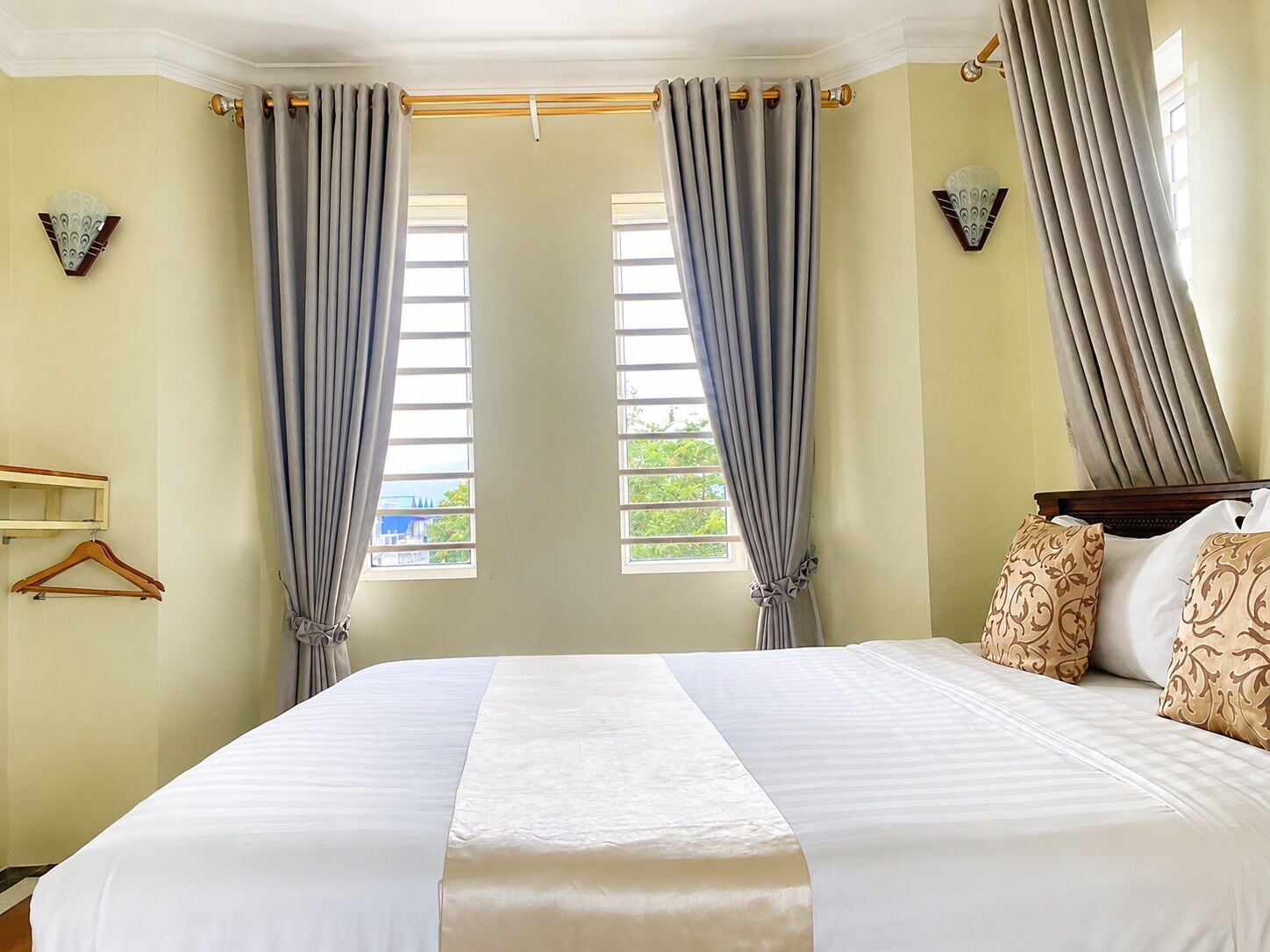 A hotel room with a large bed, two windows with curtains, a wooden headboard, and decorative wall sconces.