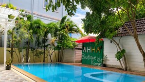 Una piscina con solárium azulejado, rodeada de vegetación y un edificio con un letrero verde.