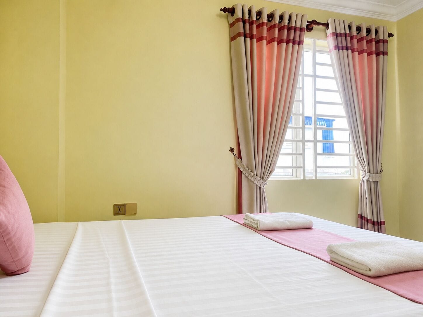 A hotel room with a bed, a window with curtains, and a wall socket.