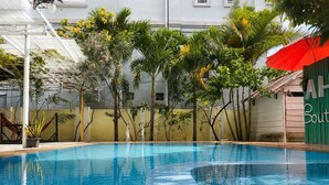 Una piscina con bordes de azulejos, rodeada de follaje y un edificio con toldo verde.