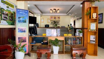 A reception area with a wooden desk, computer, and a check-in counter. There are potted plants and framed certificates on the walls.