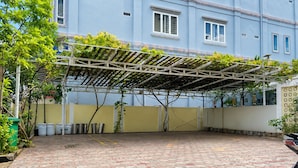 Uma área externa coberta com piso de azulejos, uma pérgola com plantas trepadeiras e um edifício ao fundo.
