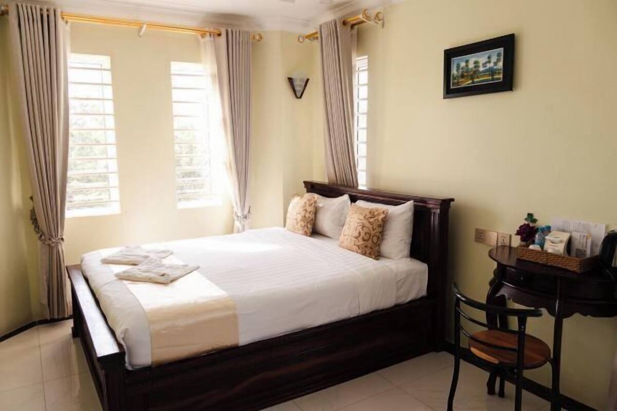 Una cama bien tendida con almohadas, un cabecero de madera, una mesita con objetos y un cuadro en la pared.