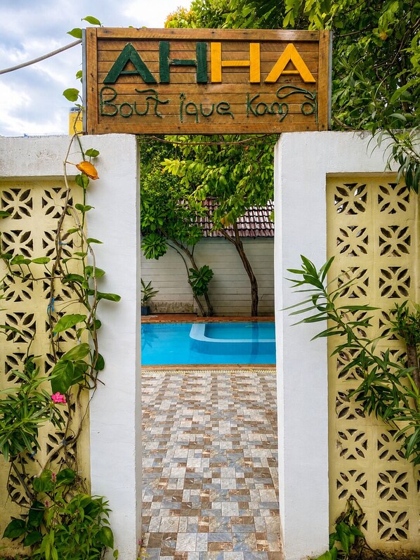 Un letrero de madera con la inscripción 'AHHA Boutique Kayak', que indica el acceso a una zona de piscina con suelo de azulejos y vistas a un jardín.