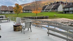 Eine Holzterrasse mit Sitzgelegenheiten im Freien, einer Steinkaminfeuerstelle und Blick auf einen Berg und Gebäude im Hintergrund.