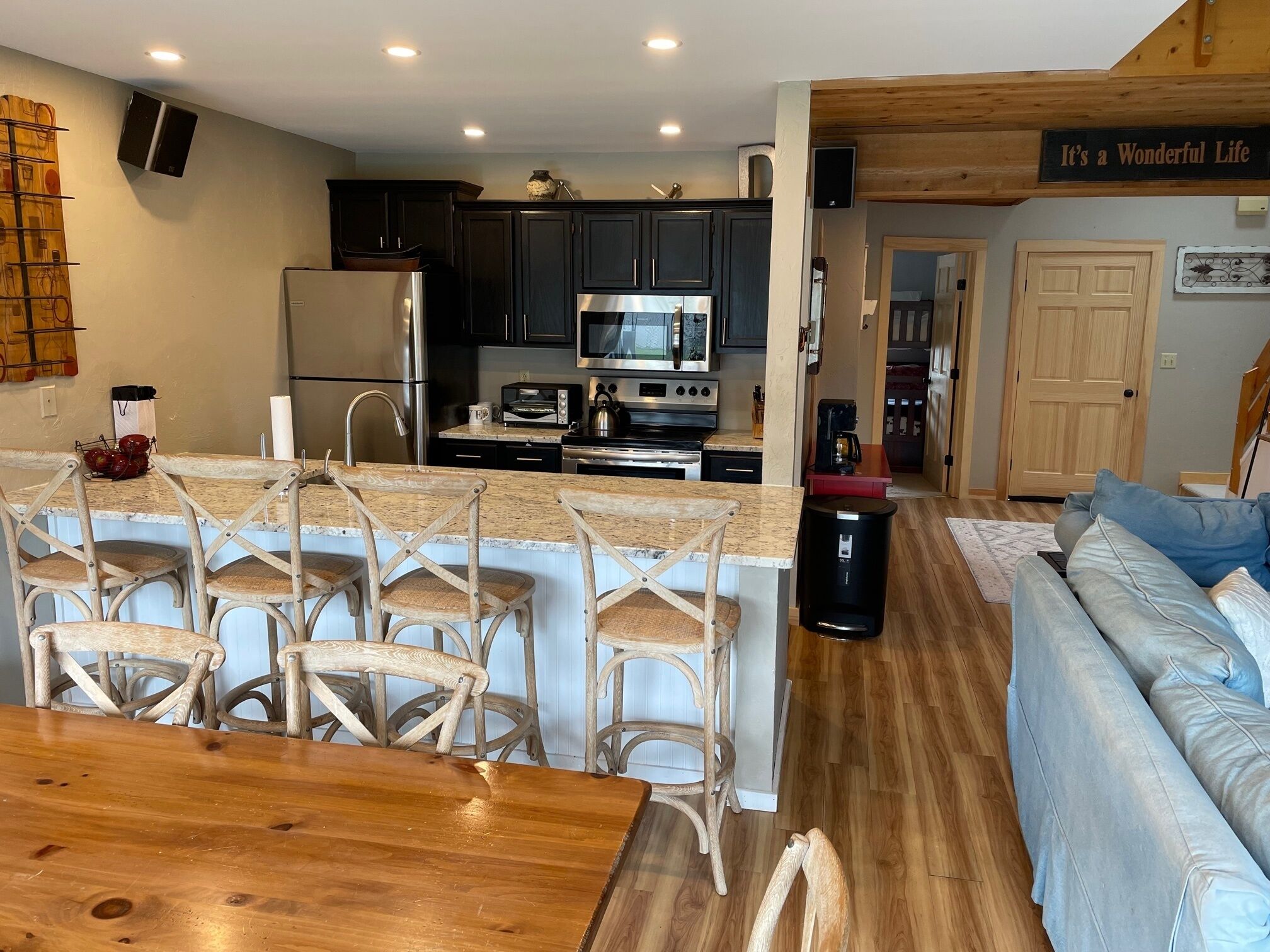 A modern kitchen with dark cabinets, a stainless steel refrigerator, a microwave, and a toaster. There is a wooden dining table with chairs and a light-colored rug in the living area.