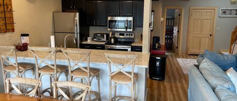 A modern kitchen with dark cabinets, a stainless steel refrigerator, a microwave, and a toaster. There is a wooden dining table with chairs and a light-colored rug in the living area.