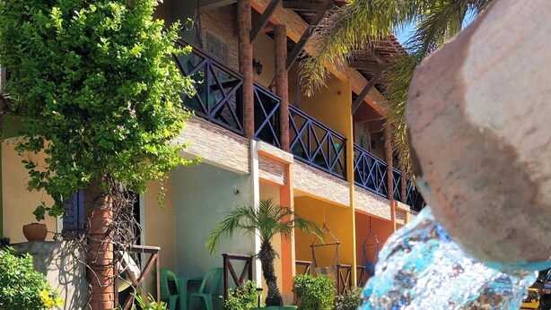 A swimming pool with clear blue water, surrounded by lush greenery and a building with a tiled roof and balconies. - Vila do Pôr do Sol  (Aracati)