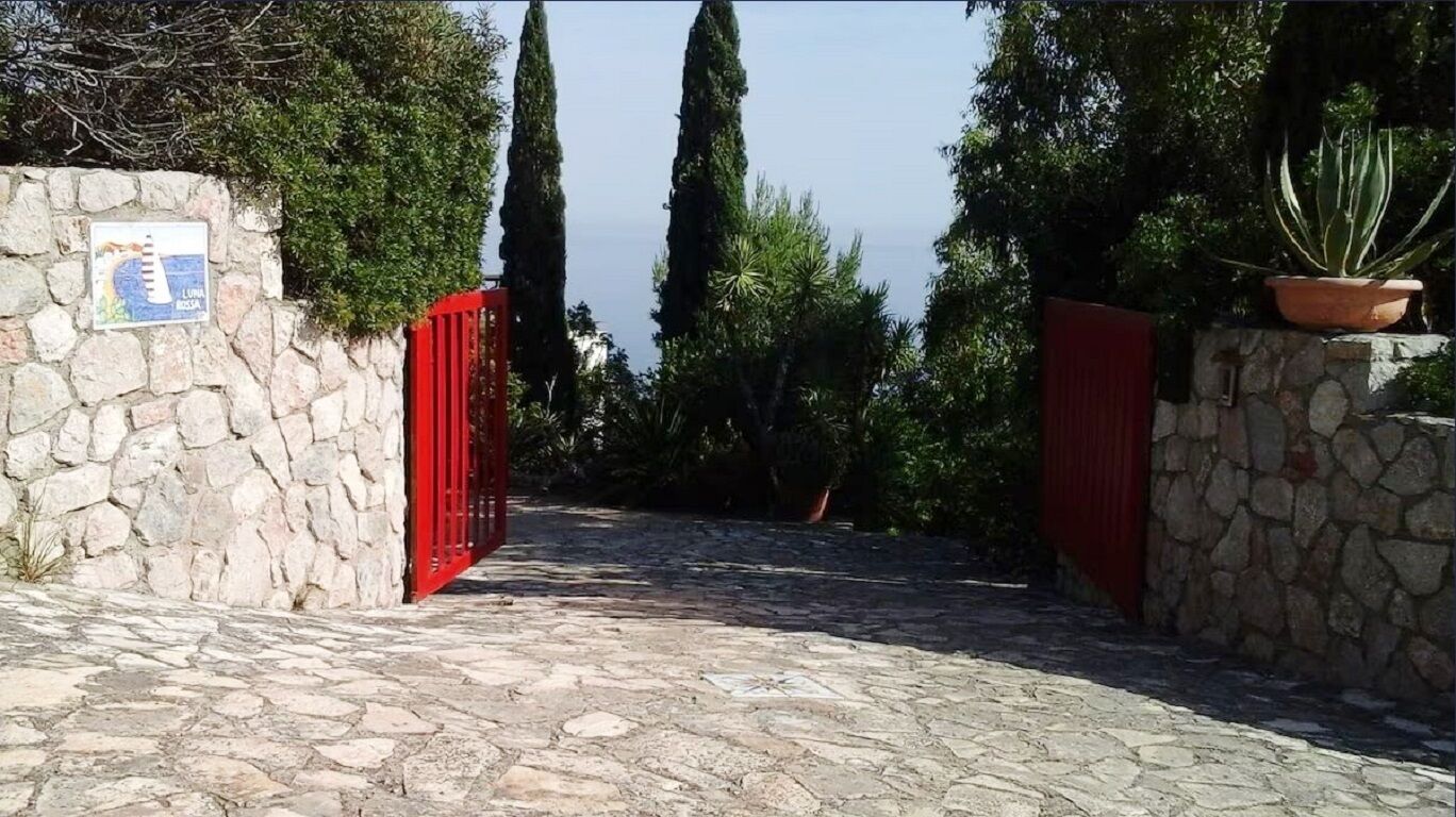 Uma trilha de pedras leva a um portão vermelho, cercado por vegetação e um muro de pedra.
