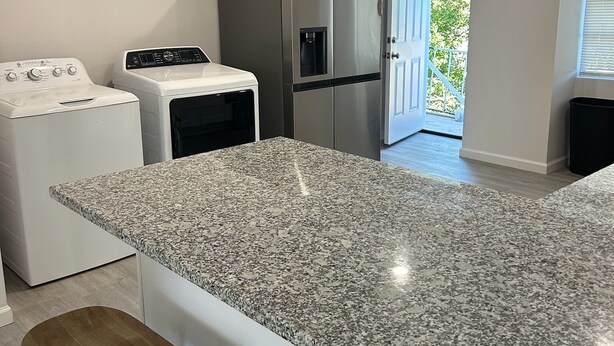 A kitchen with a granite countertop, a stainless steel refrigerator, a washing machine, and a door leading to an outdoor area. - Metairie magic, 2 bedroom gem  (Metairie)