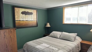 A bedroom with a bed, a nightstand, a lamp, a painting, and a window with blinds.