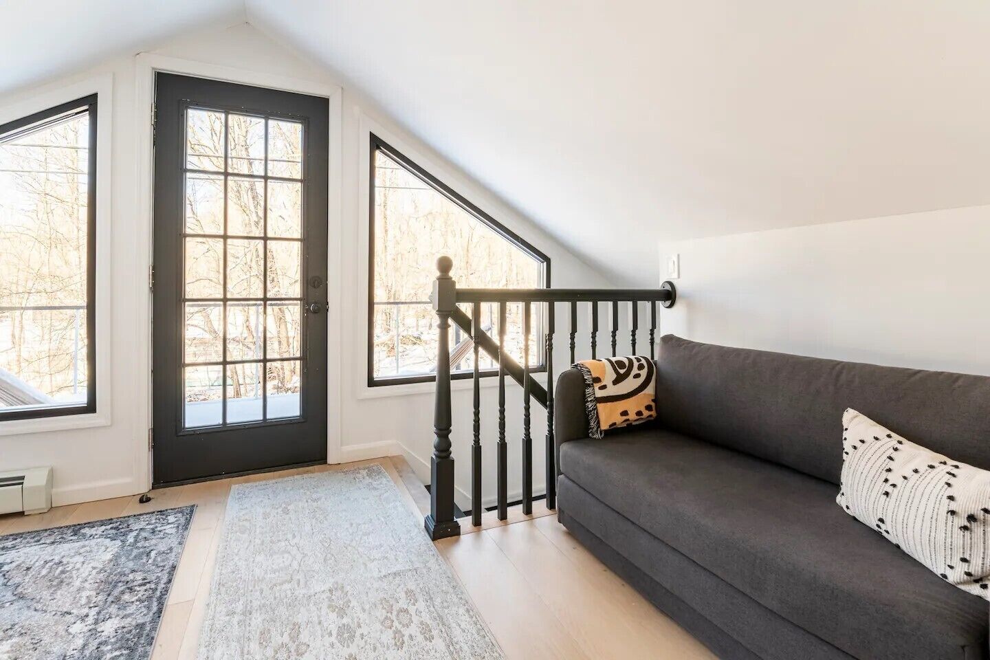 Ein Zimmer mit einem dunkelgrauen Sofa, einem gemusterten Zierkissen, einem schwarz-weißen Kissen und einem schwarzen Geländer. Ein großes Fenster bietet Blick auf Bäume.