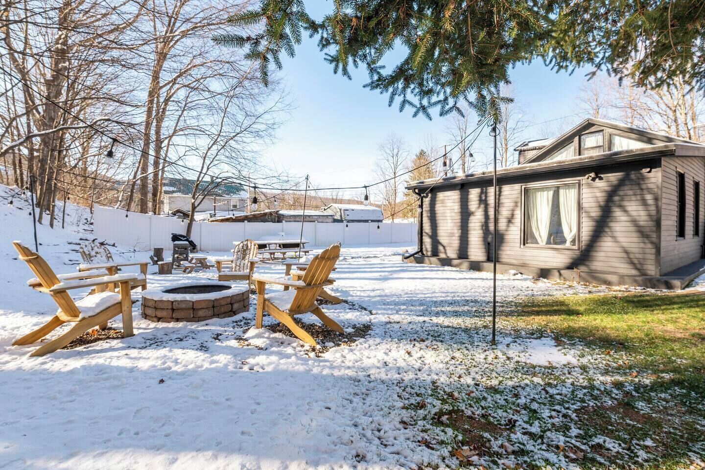 Ein mit Schnee bedeckter Garten mit Feuerstelle, Holzstühlen und einer kleinen Holzhütte.