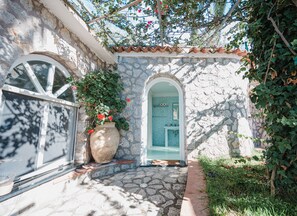 A stone building with a tiled roof, a large potted plant with red flowers, and a doorway leading to a bathroom. - Villa with Pool and 8 Double Bedrooms in Anacapri – by La Canostra Capri (Anacapri)