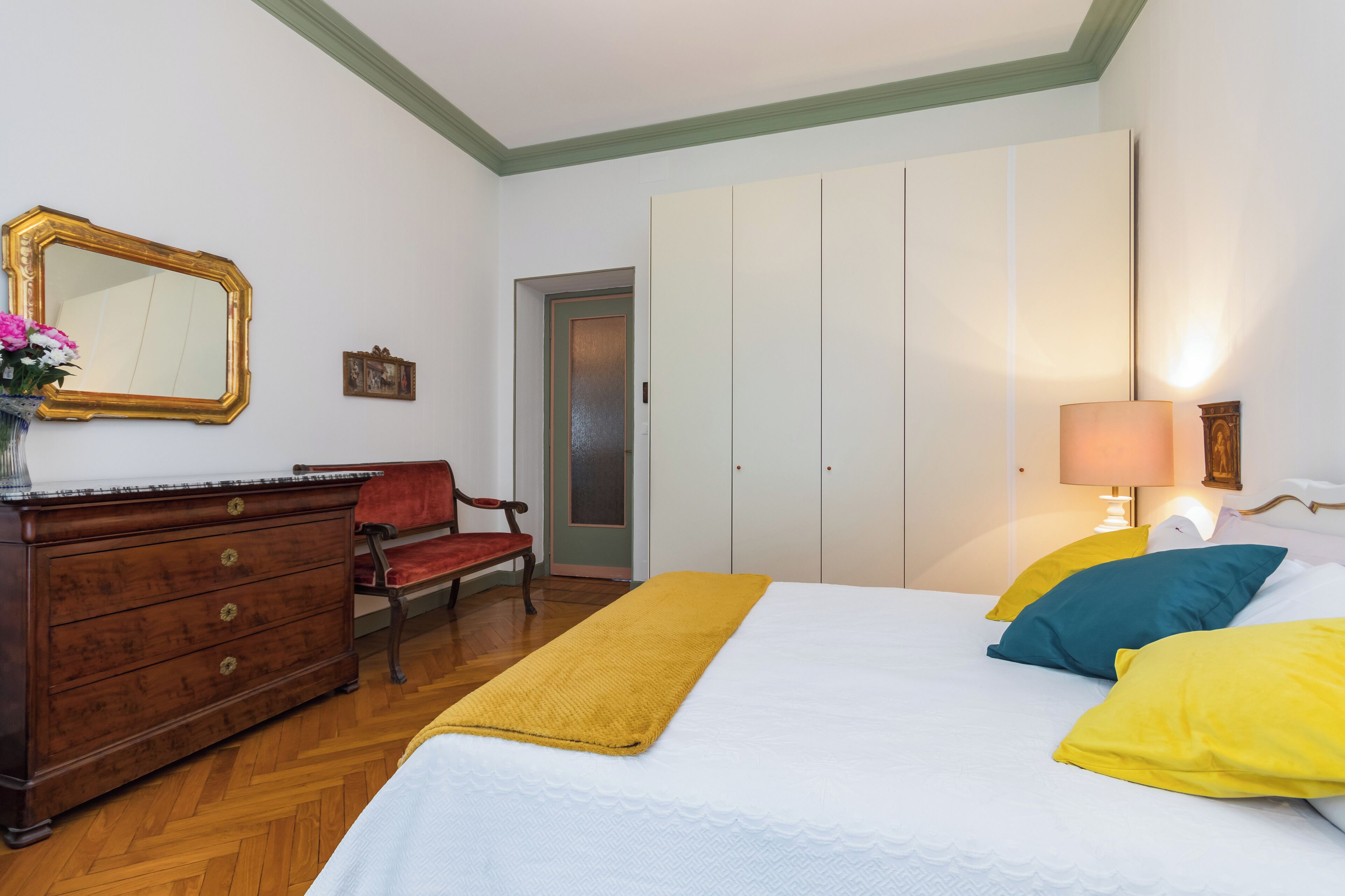 Une chambre avec une commode en bois, une chaise, un lit avec des coussins jaunes et bleus, un miroir et une porte donnant sur une autre pièce.
