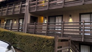 Ett hotell med flera våningar, balkonger, en parkerad bil framför och en häck längs trottoaren.