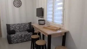 Una habitación pequeña y compacta con una zona de kitchenette que incluye una mesa de madera y cuatro taburetes. Un sofá con diseño blanco y negro está colocado contra la pared, y un reloj está colgado en la pared sobre él.