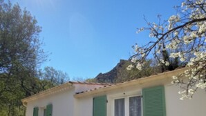 Un pequeño edificio con dos persianas verdes, fachada blanca y tejado de tejas. Hay dos sillas verdes y una pequeña mesa afuera.