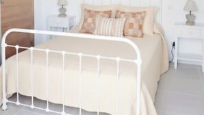 Un dormitorio con estructura de cama de metal blanco, colcha color beige y dos mesitas de noche con lámparas.