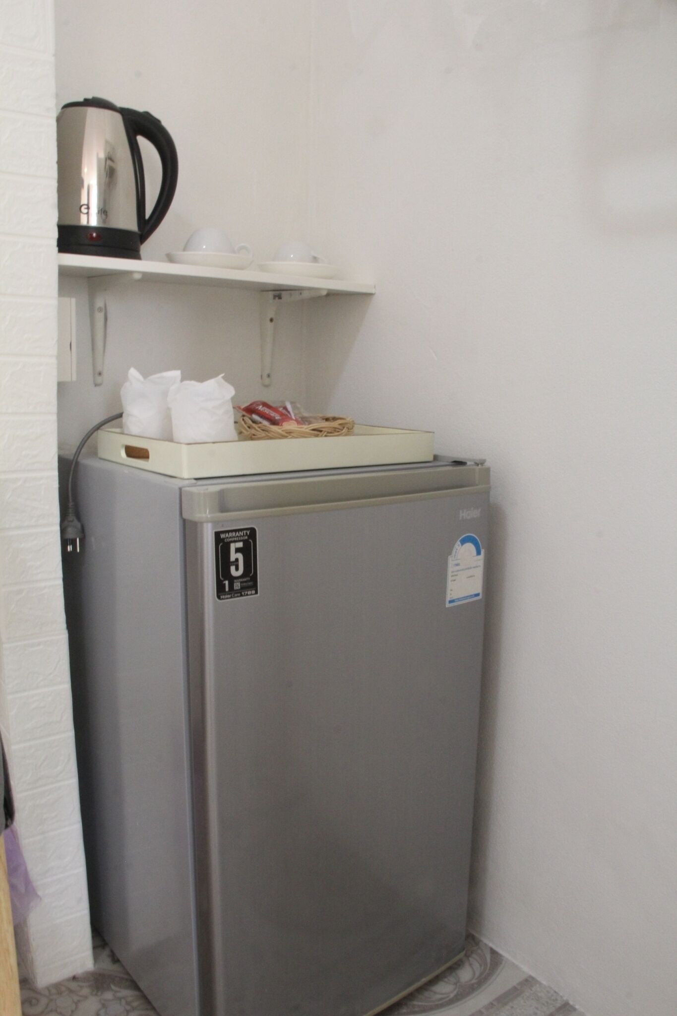 Un refrigerador compacto de acero inoxidable con un compartimento congelador, un estante pequeño encima y una tetera en la pared.