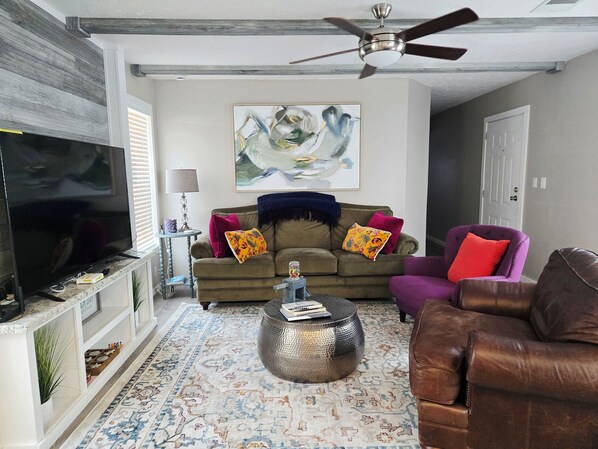 Una sala de estar con un televisor de pantalla plana, un ventilador de techo, un sofá con cojines de colores, un sillón morado y un tapete estampado.