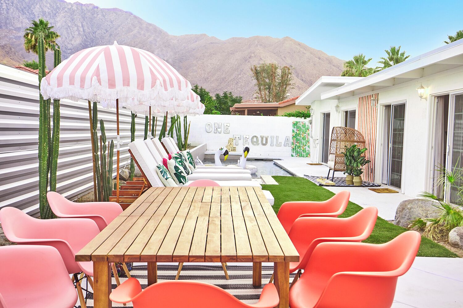 A patio with a wooden table and orange chairs, a striped umbrella, and a pool area with a sign reading "ONE TEQUILA".