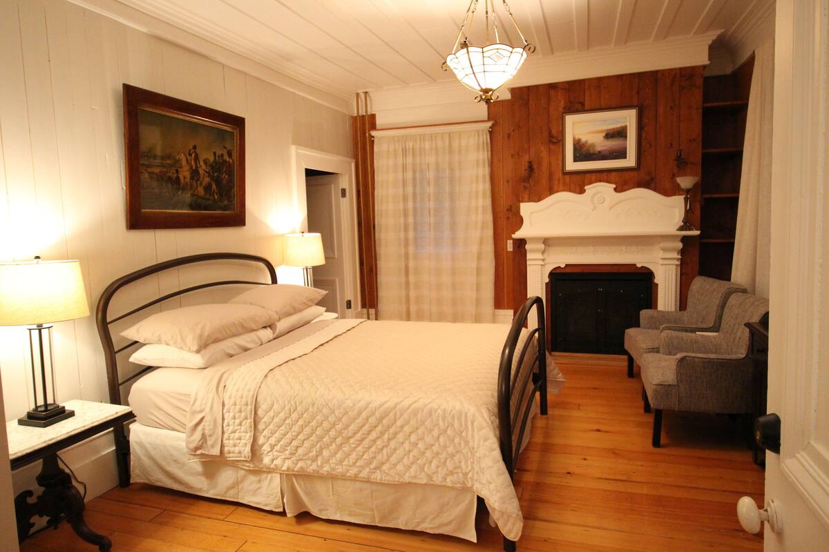 A bedroom with a bed, bedside lamps, a fireplace, and a chair.