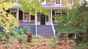 A two-story house with a front porch, steps leading to the door, and a garden with various plants and a bench. - Auberge Comme au premier jour (Saint-Pacôme)