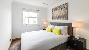 A bedroom with a large bed, a window, a wall-mounted artwork, and a nightstand with a lamp.