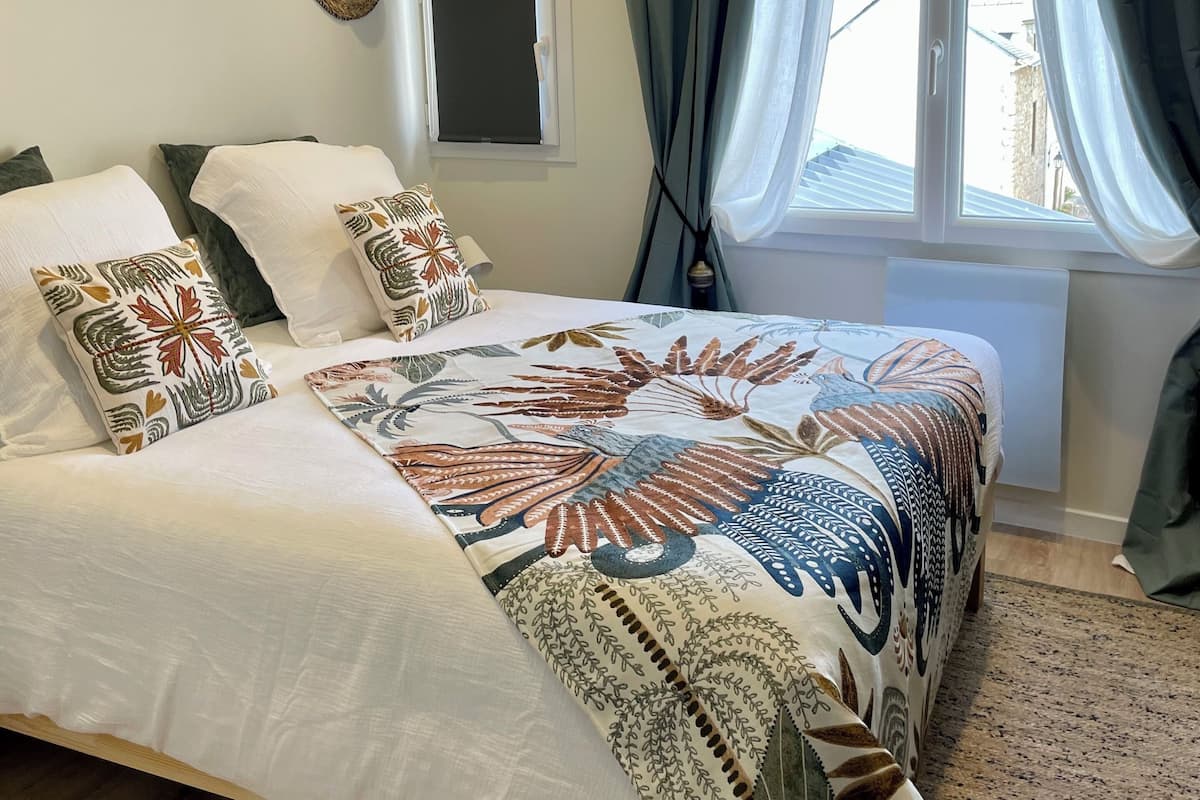 A bedroom with a bed, pillows, a window with curtains, a wall-mounted hat, and a small window with a shutter.
