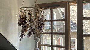 A room with a window overlooking a street, a hanging dried plant arrangement, a white textured vase, and a small decorative item on the windowsill. - Constance de Bretagne  (Batz-sur-Mer)