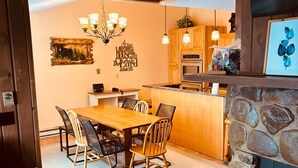 A cozy dining area with a wooden table and chairs, a stone fireplace, and a kitchen in the background. - Snowshoe Luxury Ski Lodge @ TopShelfStays (Slatyfork)