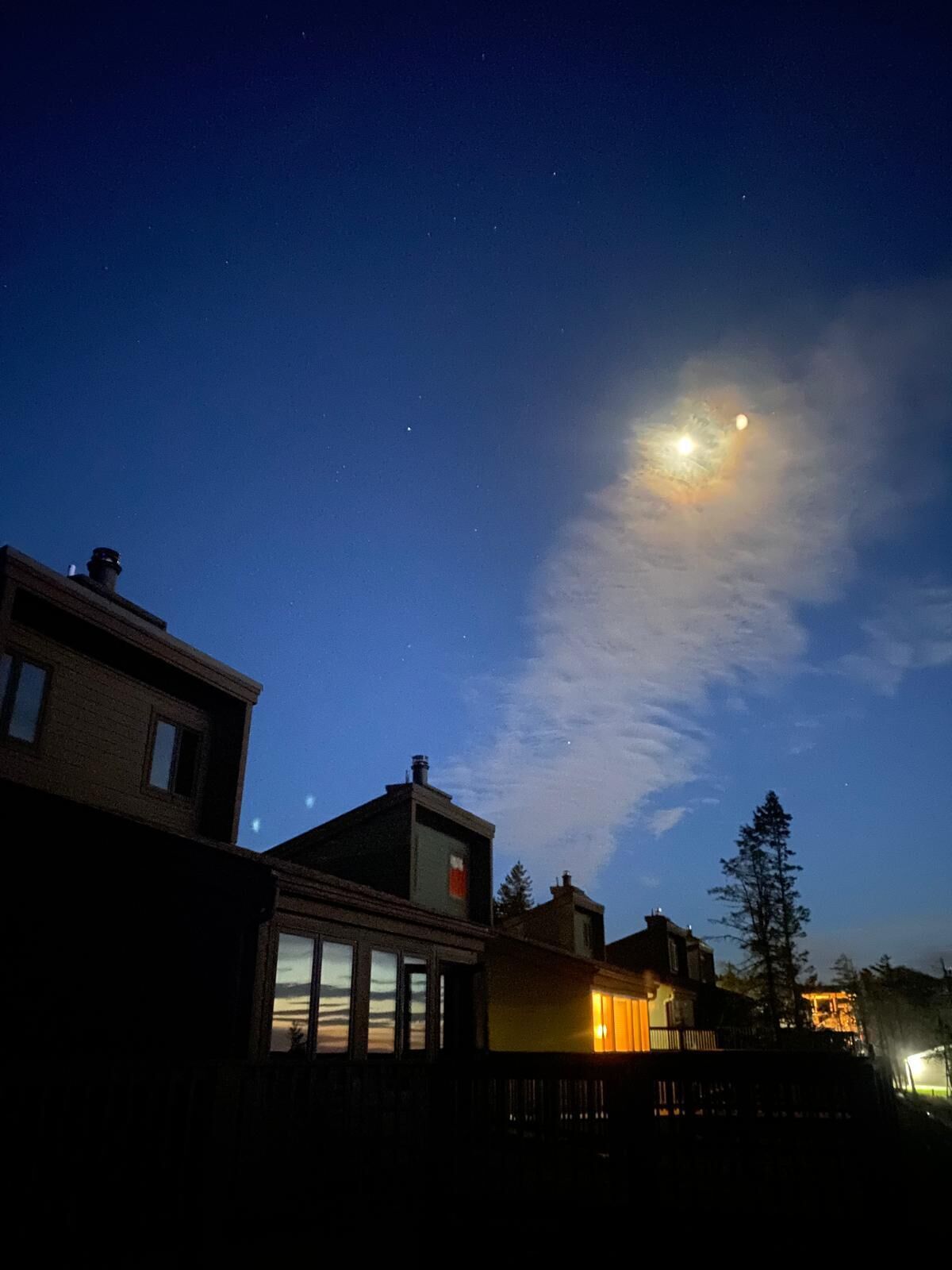 A night sky with a visible moon and stars, a wooden fence, and residential buildings with windows.