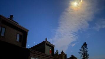 A night sky with a visible moon and stars, a wooden fence, and residential buildings with windows.