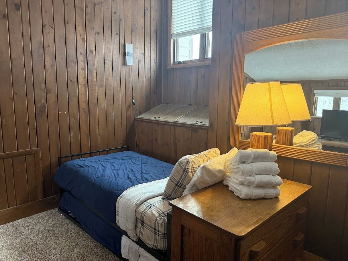 A bedroom with a wooden paneled wall, a bed with blue bedding, a wooden nightstand with a lamp, and a window with blinds.