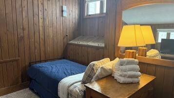 A bedroom with a wooden paneled wall, a bed with blue bedding, a wooden nightstand with a lamp, and a window with blinds.