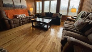 A cozy living room with wooden paneling, two leather sofas, a coffee table, and large windows with blinds. - Snowshoe Luxury Ski Lodge @ TopShelfStays (Slatyfork)