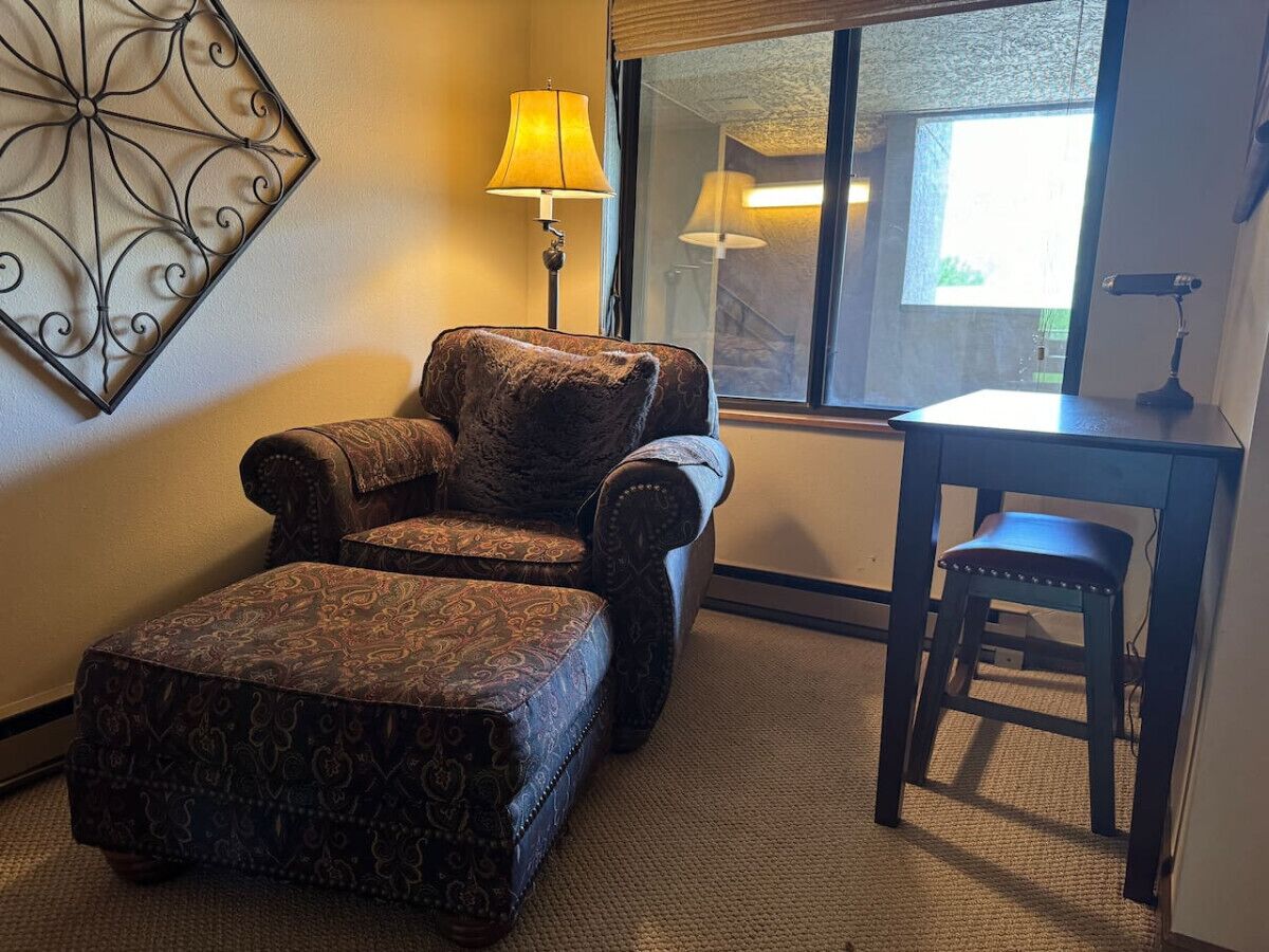 A room with a patterned armchair and ottoman, a small table, a lamp, and a view through a window.