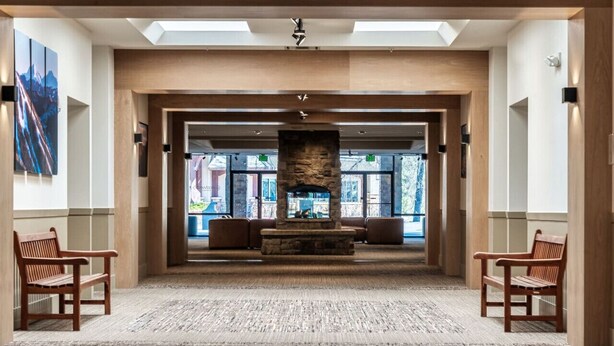 Un hall d’entrée spacieux avec une cheminée en pierre, des bancs en bois et une vue sur l’extérieur grâce à de grandes fenêtres.