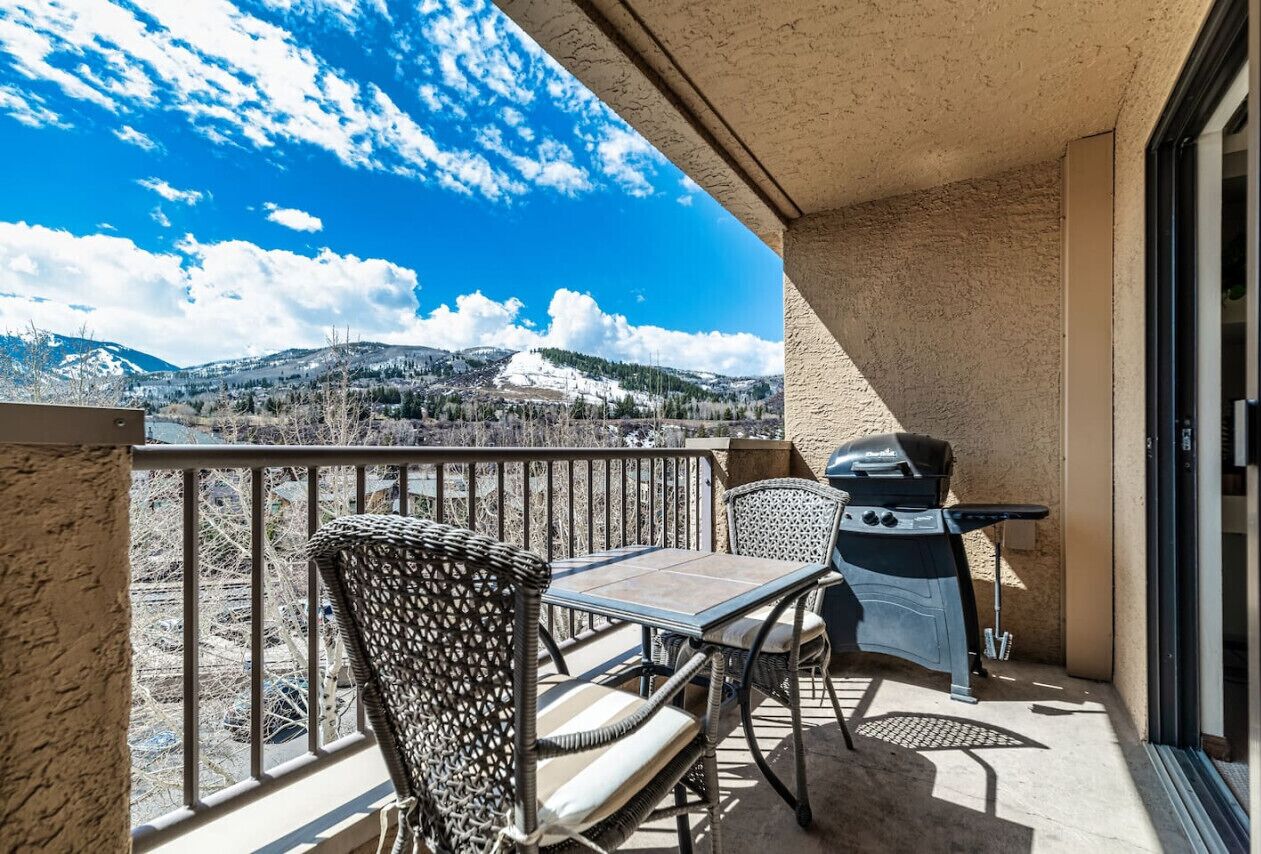 A balcony with a view of snow-covered mountains, a grill, and wicker furniture.