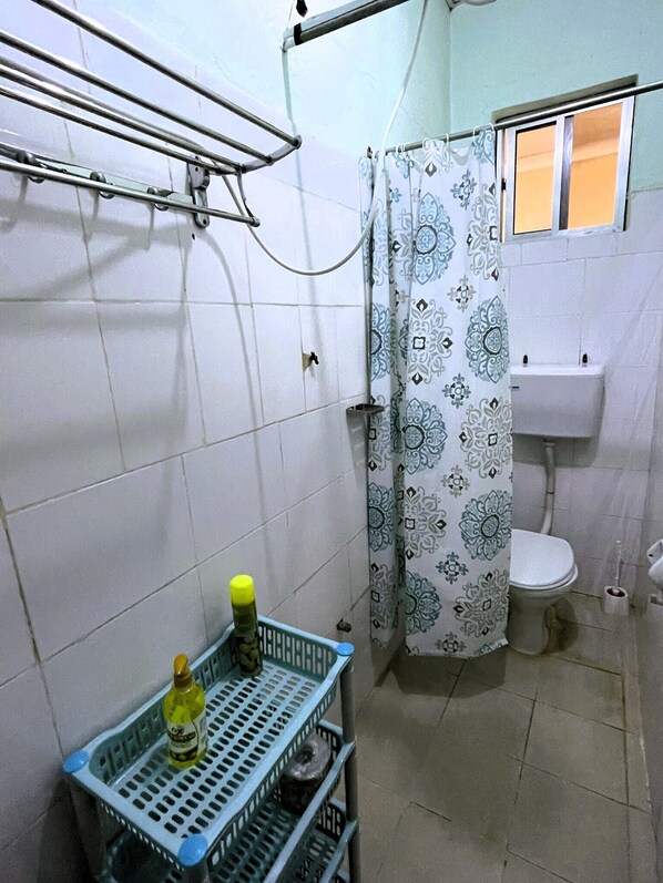 Un baño pequeño con inodoro, lavamanos, cortina de ducha y un estante con productos de limpieza.
