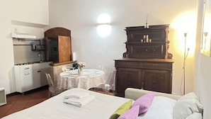 Un dormitorio con cama, mesitas de noche, cómoda, una pequeña mesa con sillas y un área de cocina con estufa y fregadero.