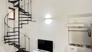 Un salón con una escalera espiral, un televisor de pantalla plana sobre un mueble blanco, una zona de cocina con estufa y un reloj en la pared.