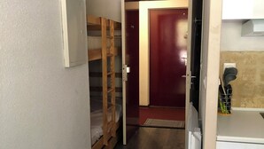 Un couloir étroit avec un lit superposé, une porte, un réfrigérateur et un coin cuisine équipé d’un évier et d’ustensiles.