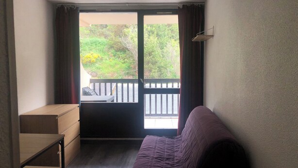 Une petite pièce avec un canapé violet, une table en bois et une porte coulissante donnant sur un balcon d’où l’on peut admirer la verdure.
