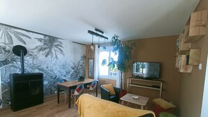 A living room with a wooden floor, a sofa with a yellow blanket, a dining table with chairs, a TV, and a mural of palm trees. - Maison Avec Jardin Proche Centre Ville, 15 min de la Plage, 25 min de Brest (Lesneven)