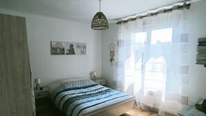 A bedroom with a wooden floor, a bed with patterned bedding, a hanging light fixture, a bedside table with a lamp, and a window with sheer curtains. - Maison Avec Jardin Proche Centre Ville, 15 min de la Plage, 25 min de Brest (Lesneven)