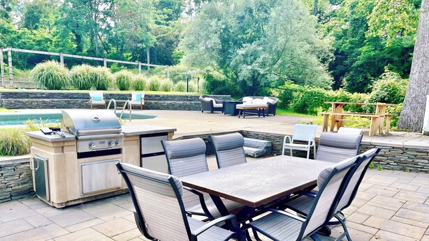 An outdoor patio with a stone table and chairs, a grill, and a pool in the background. - Cape Cod Estate: 2 Homes, Pool, Tennis Court, Game Barn and Bar, Sleeps 20 (Sandwich)